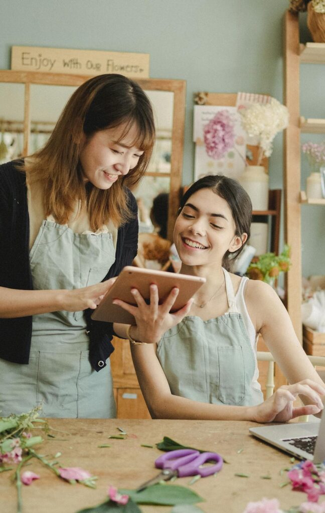 Content multiethnic female florists in aprons at table with laptop and browsing tablet while preparing for order in creative floristry studio